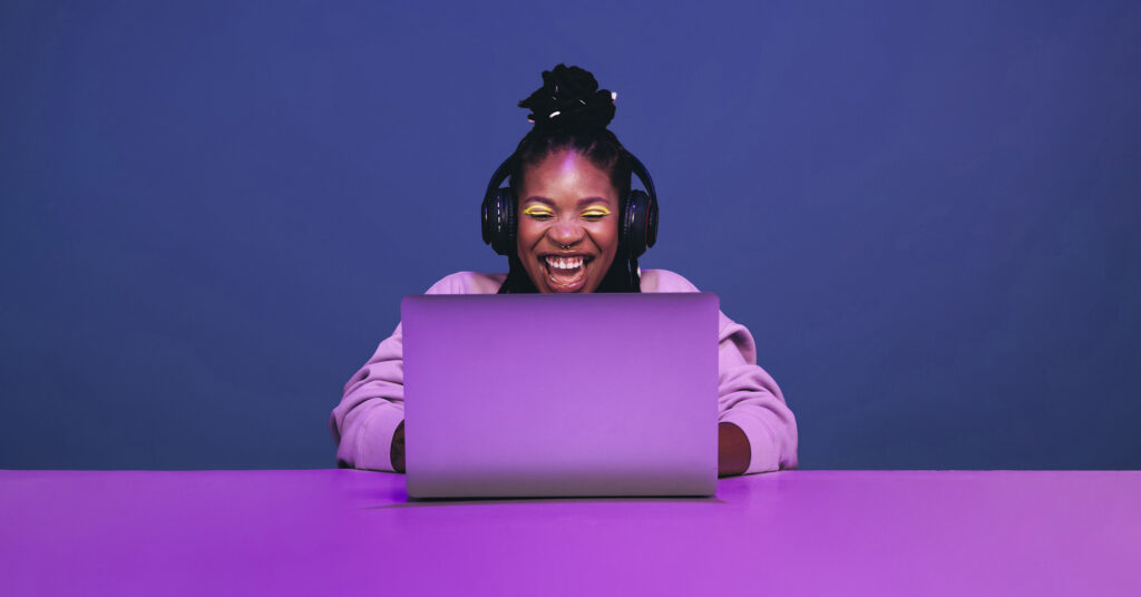 Woman with makeup, piercings and headphones laughing at laptop from face