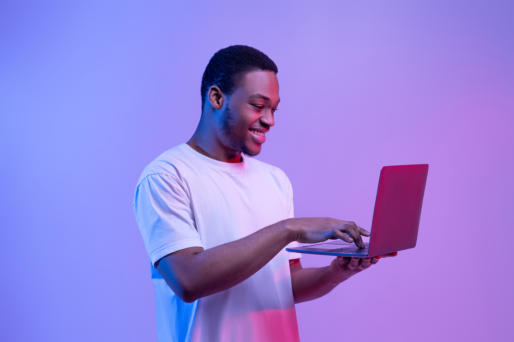 Man standing, smiling and typing on laptop. Purple background light.